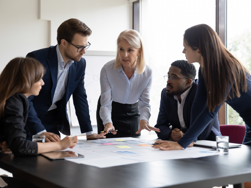 Fünf Geschäftsleute diskutieren Strategien und Dokumente am Konferenztisch im Büro und entwickeln gemeinsam innovative Individuallösungen für komplexe Herausforderungen.