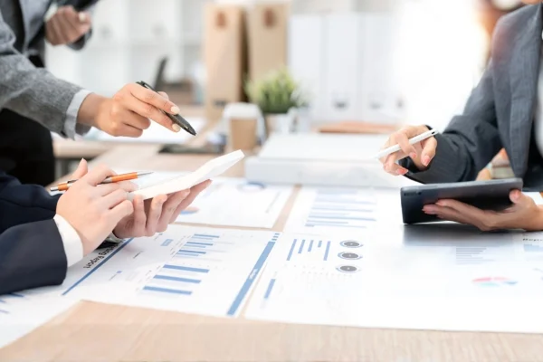 Mehrere Geschäftsleute analysieren Diagramme und Daten am Tisch mit Tablet und Unterlagen und entwickeln gemeinsam passende Individuallösungen.